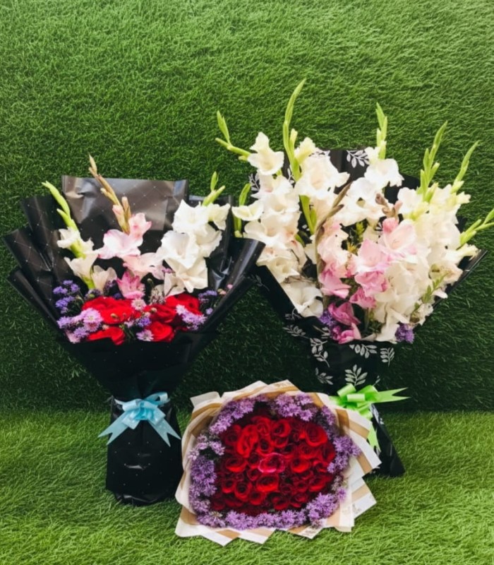 Three premium mixed flower bouquets with red roses, white and pink gladiolus, and colorful filler flowers, beautifully wrapped with ribbons, displayed on artificial green turf – Timeless Wrap Gift Shop